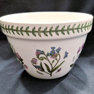 May include: A white ceramic mixing bowl with a green leaf pattern around the rim and floral and butterfly designs on the sides. The bowl has a wide, rounded shape and is suitable for kitchen use.