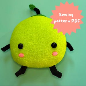 Peut inclure: Un jouet en peluche vert lime avec des jambes noires et une feuille verte sur le dessus. Le jouet a deux yeux noirs et deux joues roses. Le texte "Sewing pattern PDF" est dans un éclat d'étoile rose.