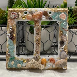 May include: A white, square light switch cover decorated with various seashells, pearls, and gold accents. The frame is embellished with small pieces of sea glass and coral, creating a coastal theme. The cover has two switch openings.