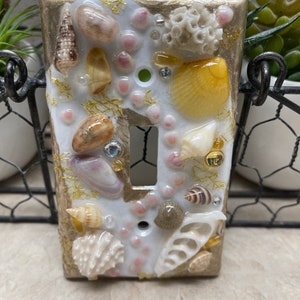 May include: Decorative light switch cover featuring a coastal theme. The rectangular cover is adorned with various seashells, coral, and small beads, set against a white and gold background. The switch plate has a toggle switch opening and screw holes.