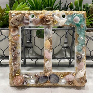 May include: A decorative light switch cover adorned with seashells, gold accents, and clear resin. The frame is rectangular with a double switch opening. Various seashells and small embellishments are embedded in the resin, creating a coastal theme.