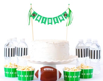 May include: A cake topper banner with green pennants spelling AIDEN, flanked by two football icons on wooden sticks. Below sits a white frosted cake on a stand, a brown football, four water bottles with black and white striped labels, and four green popcorn cups with white yard line markings.