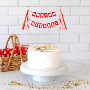 May include: A round cake with white frosting and colorful sprinkles, topped with two wooden sticks holding two red pennant banners. The top banner reads CUSTOM and the bottom banner reads MESSAGE in white letters. Red ribbons hang from the sticks.
