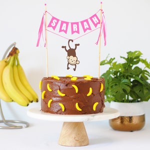 May include: A chocolate cake decorated with yellow banana candies. A pink banner with the name "SARAH" is displayed above the cake, along with a brown monkey illustration. The cake sits on a white marble cake stand.