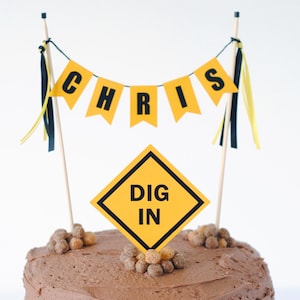 May include: A chocolate frosted cake with a banner that says "CHRIS" and a yellow and black "DIG IN" sign. The cake is decorated with small, round candies and is topped with wooden skewers.