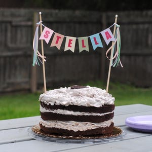 May include: A layered chocolate cake with white frosting and a banner that reads "STELLA". The banner is attached to two wooden sticks and has colorful flags. The cake sits on a gold-colored plate.