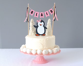 May include: A cake topper set featuring a penguin with a pink bow, two white textured trees, and a pink banner with the name BLAIR in black letters. The set includes two wooden sticks with pink and black ribbons attached to the top ends.