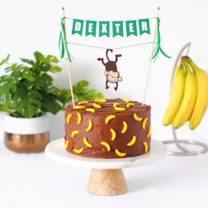 May include: A chocolate cake decorated with yellow banana-shaped candies. A banner with the name "DEXTER" is suspended above the cake, along with a cartoon monkey. The cake sits on a white cake stand with a wooden base.