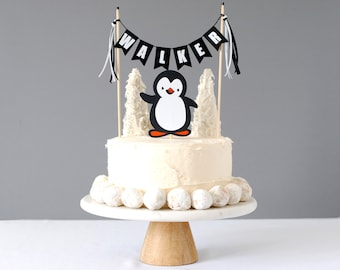 May include: A cake topper set featuring a black and white penguin illustration, two white textured tree-shaped cutouts, and a black bunting banner with white letters spelling WALKER, all attached to wooden sticks with black and white ribbons.