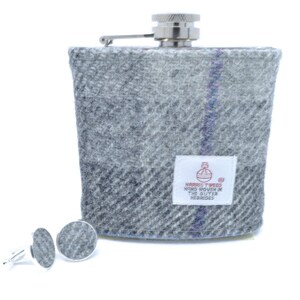 May include: A metal flask covered in grey and white plaid wool fabric with a single thin blue vertical stripe. A white rectangular label features the text Harris Tweed Hand Woven In The Outer Hebrides. Two matching circular wool cufflinks are included.