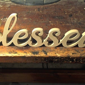 May include: A wooden sign with the word "Blessed" in a cursive font. The sign is made of light brown wood and is sitting on a wooden surface.