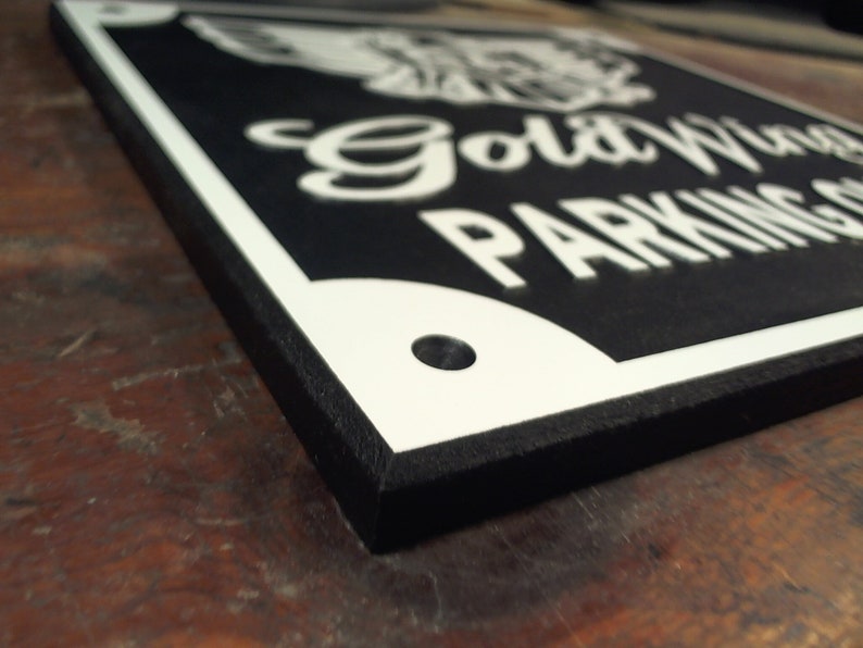 Peut inclure: Un panneau noir et blanc avec un motif d'aigle blanc et le texte "Gold Wing Parking Only". Le panneau a une bordure noire et un trou dans le coin sup&eacute;rieur droit.