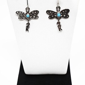 May include: Sterling silver dragonfly earrings with turquoise stones and 2K markings. The earrings are displayed on a white stand with a black base.