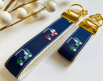 May include: Two navy blue and white keychains with gold key rings. The keychains feature a repeating pattern of colorful golf carts.