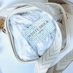 May include: Cream-colored crossbody bag with a patterned strap. The bag features a rectangular shape and a clear front. Inside the bag is white tissue paper and a card that reads "WHITE BRICK GIFTS."