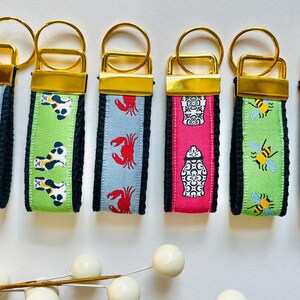 May include: Seven colorful keychains with gold key rings. Each keychain has a different design, including a palm tree, a dog, a crab, a jar, a bee, and a coffee cup.