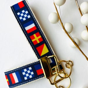 May include: A navy blue and cream-colored keychain strap with nautical flag designs in red, white, blue, and yellow. The strap is attached to a gold-colored metal clasp and key rings.