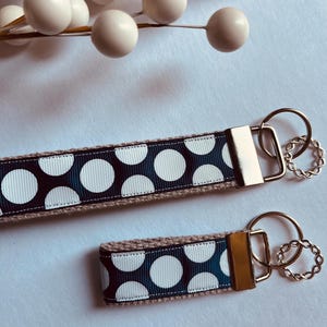 May include: Two keychains with a navy blue and white polka dot pattern. The larger keychain has a silver key ring and the smaller keychain has a gold key ring.