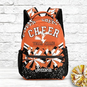 Puede incluir: Mochila naranja y negra con la palabra "CHEER" en letras blancas. La mochila presenta un gráfico de animadora, un diseño de pompones y el nombre "Jessica". La mochila tiene bolsillos laterales de malla negra y correas negras.