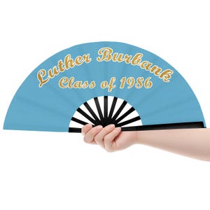 May include: A light blue folding hand fan with black ribs and the gold-coloured text Luther Burbank Class of 1986 printed in a cursive and block font across the top half of the fan surface, creating a commemorative design.