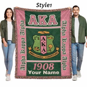 Op de afbeelding: Een roze en groene geweven deken met de tekst "AKA" en "Alpha Kappa Alpha". De deken heeft een wapen en het jaar "1908". De woorden "Your Name" worden ook weergegeven.