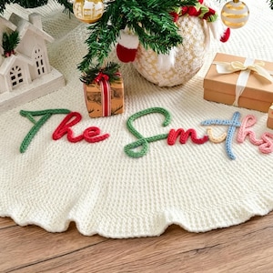 May include: A circular, off-white knitted Christmas tree skirt featuring the text The Smiths embroidered in a multi-colored yarn script. The letters are crafted in shades of green, red, light blue, yellow, and pink, creating a decorative accent.
