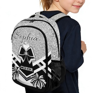 May include: A silver glitter backpack with a black and white cheerleading design. The backpack features the name "Sophia" in script, a bow, pom-poms, and the word "CHEER". It has a black mesh side pocket and black straps.