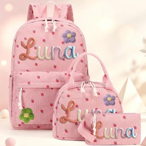 May include: A pink corduroy backpack, lunch bag, and pencil case set featuring a repeating strawberry pattern. Each item is personalized with the name Luna in multi-colored yarn lettering and decorated with small, textured fabric flower appliques.