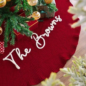 May include: A circular, deep red knitted Christmas tree skirt featuring the white embroidered text "The Browns" in a cursive script. The skirt is positioned under a green pine tree decorated with gold ornaments and wrapped gift boxes.