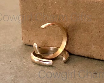 Könnte beinhalten: Zwei goldene Ohrringe, einer auf dem anderen, auf einer braunen Oberfläche. Die Ohrringe sind aus dünnem, flexiblem Metall gefertigt und für das Tragen am oberen Teil des Ohrs konzipiert.