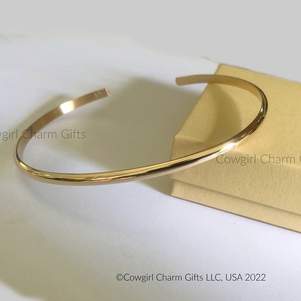 May include: A gold-toned bangle bracelet with a smooth, rounded surface. The bracelet is displayed on a light brown box with the text "Cowgirl Charm Gifts" printed on it.