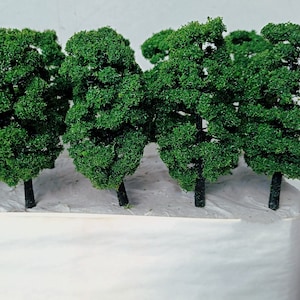 May include: A group of miniature model trees with dark green foliage and black trunks. The trees are arranged in a row, with a white base beneath them. The trees are designed for use in model train sets, dioramas, or other miniature scenes.