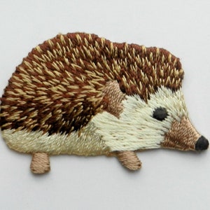 Könnte beinhalten: Gestickter Igel-Aufnäher mit detailliertem Design. Der Igel hat einen braunen und beigefarbenen stacheligen Rücken, einen cremefarbenen Bauch und eine schwarze Nase und Augen. Der Aufnäher ist ein niedliches, skurriles Design.