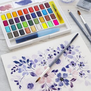 May include: A watercolor painting setup featuring a white palette with an array of colorful paint blocks, a water brush, and paintbrushes. A watercolor floral painting in shades of purple and pink is in progress.