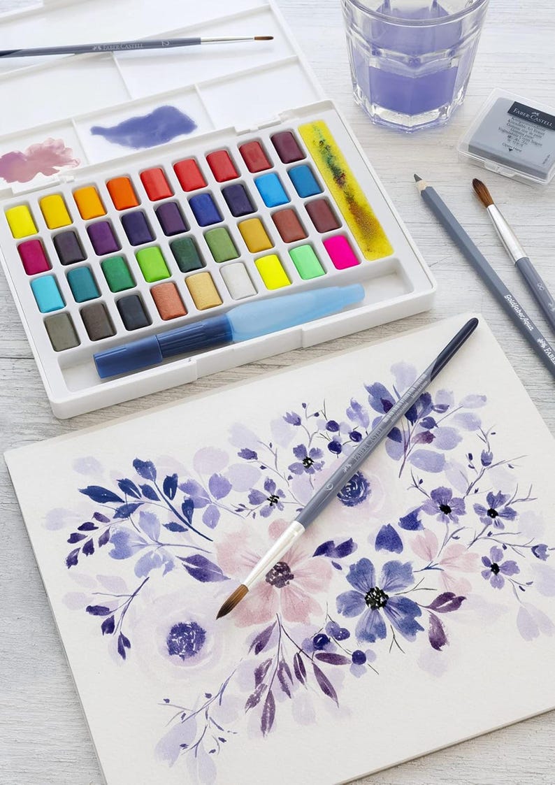 May include: A watercolor painting setup featuring a white palette with an array of colorful paint blocks, a water brush, and paintbrushes. A watercolor floral painting in shades of purple and pink is in progress.
