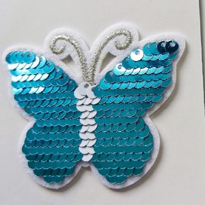 May include: A turquoise blue butterfly applique with white sequins. The butterfly is made of fabric and has a white felt backing.