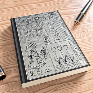May include: A hardcover notebook with a decorative silver cover featuring the words "GARDEN NOTES" and various garden-themed illustrations. A black pen with gold accents rests on the wooden surface next to the notebook.