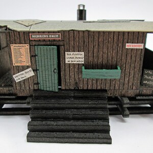May include: A detailed miniature model of a rustic wooden shack. The structure features a weathered appearance with a green door, a small green planter, and several signs, including "Members Only" and "Coal Jacks Wanted".