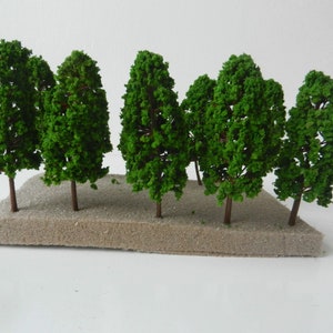May include: A miniature model of a grove of trees. The trees have green foliage and brown trunks, set on a tan base. The trees are arranged in a cluster, creating a small forest scene.