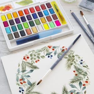 May include: A watercolor painting set with a variety of vibrant colors, a water brush, and paintbrushes. A watercolor painting of a wreath with berries and leaves is in progress. A glass of green liquid is also present.