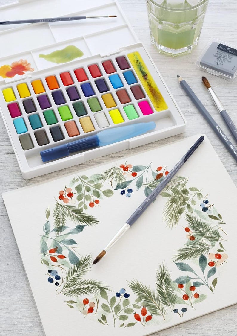 May include: A watercolor painting set with a variety of vibrant colors, a water brush, and paintbrushes. A watercolor painting of a wreath with berries and leaves is in progress. A glass of green liquid is also present.