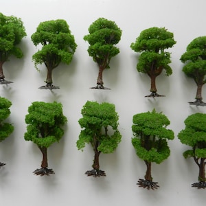 May include: Ten miniature model trees are displayed. Each tree features a brown trunk, vibrant green foliage, and a dark base. The trees are arranged in two rows against a white backdrop. These trees are suitable for model railway layouts or dioramas.