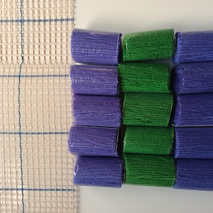 May include: An overhead shot shows a collection of wrapped yarn bundles in shades of purple and green. The bundles are arranged in rows, with a woven rug canvas visible on the left. The yarn is likely for rug hooking or punch needle projects.