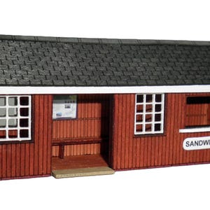 May include: A miniature red cafe with a dark gray shingled roof. The building has white-framed windows and a small bench. A sign reads "Sandwiches" and another says "Cafe".