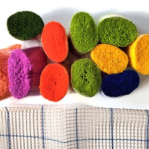 May include: A selection of colourful rug-making materials. Bundles of bright yarn in shades of green, orange, pink, yellow, and blue are arranged on a white surface. A rug canvas with a grid pattern is visible at the bottom.