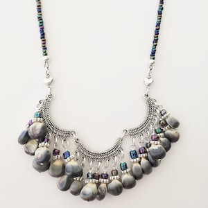 May include: A silver necklace with a crescent moon pendant and a fringe of gray seed pods. The necklace is accented with iridescent beads.