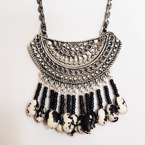May include: A silver necklace with a large, ornate pendant and black and white seed beads. The pendant is shaped like a crescent moon and has a detailed design of chains and circles. The beads are strung on thin black cords and hang from the bottom of the pendant.