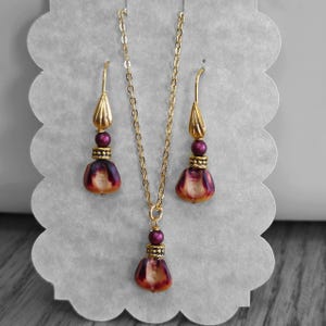 May include: A jewelry set consisting of a necklace and two drop earrings. Each piece features a teardrop-shaped bead with a gradient of deep purple to cream, a small metallic spacer, a dark purple round bead, and a gold-toned metal component.