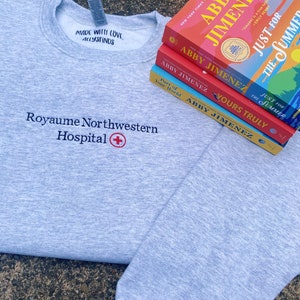 Könnte beinhalten: Ein graues Sweatshirt mit dem Text "Royaume Northwestern Hospital" und einem roten Kreuz, das auf der Vorderseite gestickt ist. Das Sweatshirt liegt auf einer braunen Oberfläche mit einem Stapel von vier Büchern darauf. Die Bücher sind alle von der gleichen Autorin, Abby Jimenez, und haben bunte Umschläge.