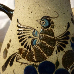 Puede incluir: Una jarra de cerámica con un diseño de pájaro marrón y azul. El pájaro tiene una cola larga y está posado sobre una flor azul. La jarra está decorada con un fondo blanco y detalles marrones y azules.